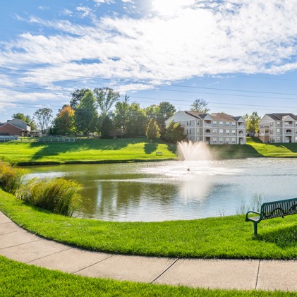 Concrete walkway in front of a n outdoor pond with a fountain in the middle of grass in front of Columns on Wetherington apartments in Florence, KY